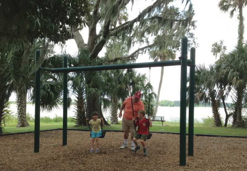 Man in orange pushing a boy in red on a swing while girl in yellow and blue swings on the side with lake and trees behind them.