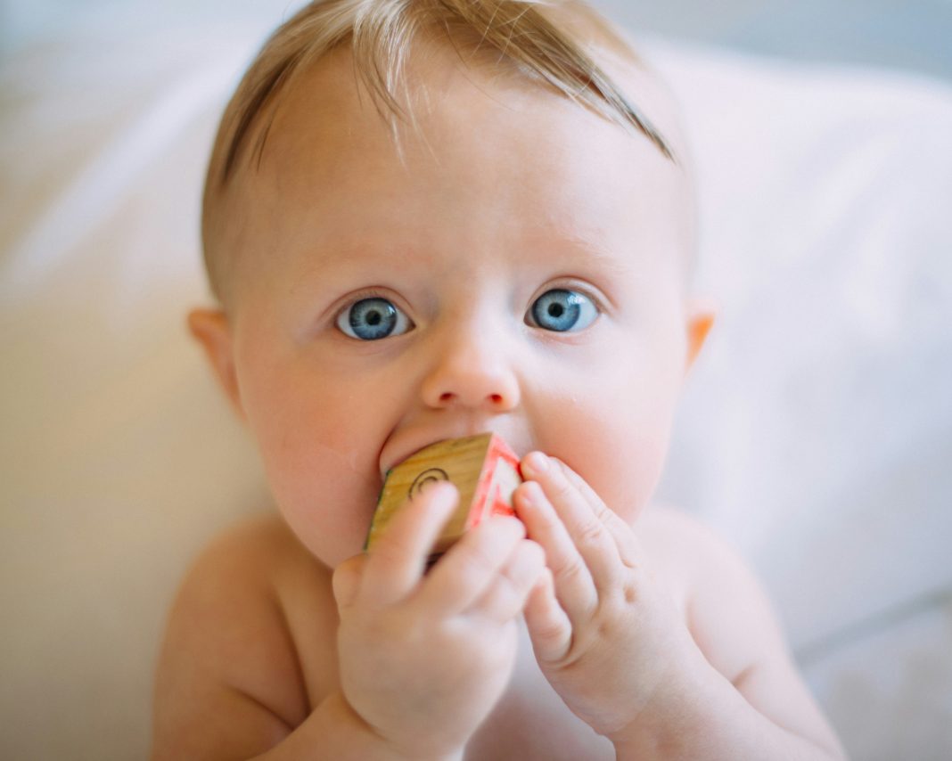 colin-maynard via unsplash baby with blonde hair and blue eyes on a white blanket with a block in his mouth