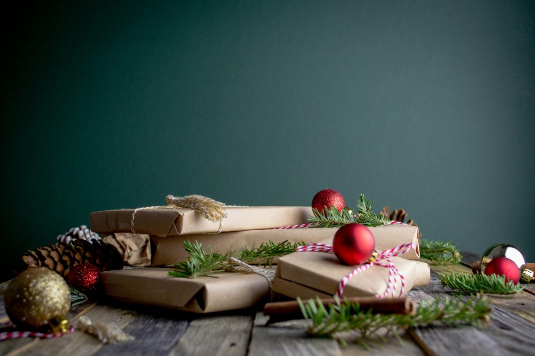 mel-poole via unsplash presents wrapped in brown paper with Christmas tree branches and ornaments around on a dark gray background and a gray wood table