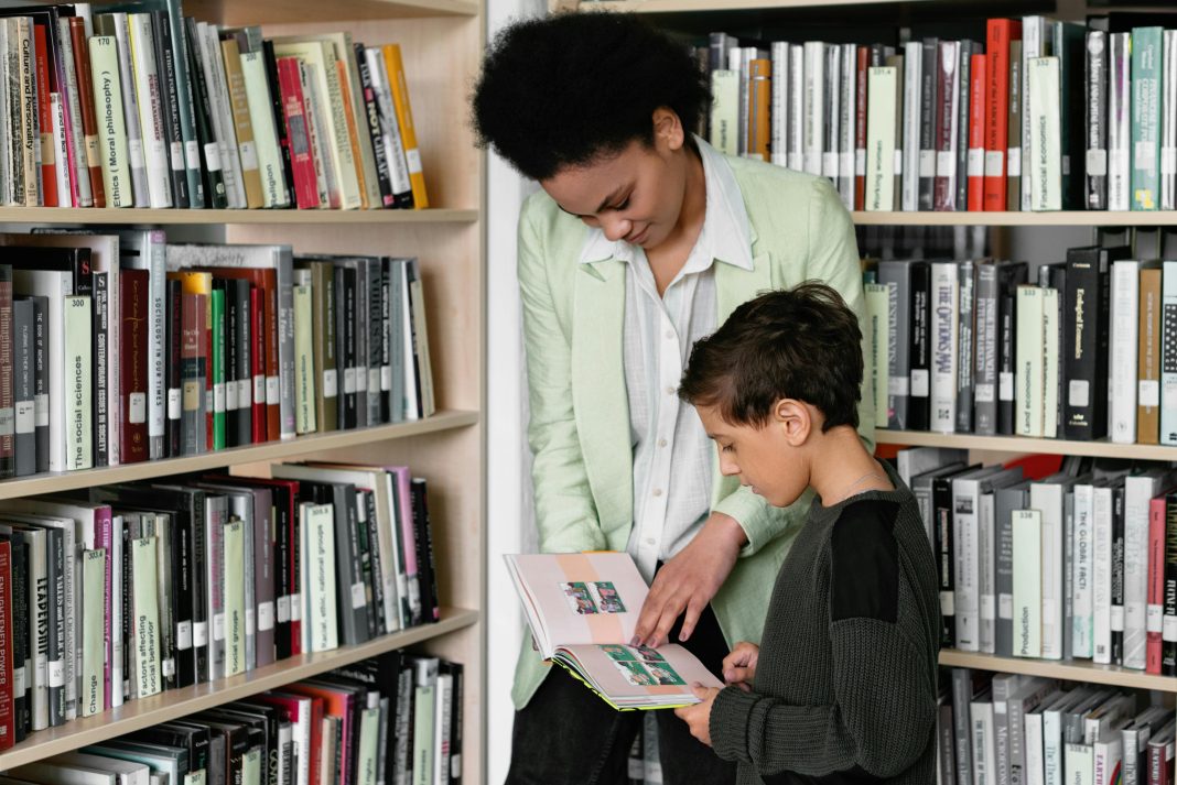 PTA mom guiding a young boy through book choices in a bright school library aisle.