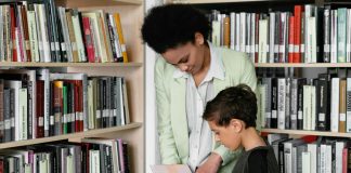 PTA mom guiding a young boy through book choices in a bright school library aisle.