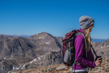 woman with purple coat and gray hat on, backpacking with mountains behind her