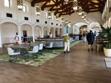 inside of hotel with wood floor and green carpet, lights hanging in rows from the ceiling and people walking around