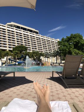 woman's feet on a pool chair with a pool, fountain and hotel behind