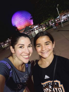 Mom and daughter smiling with Epcot Spaceship Earth lit up in shades of purple behind them