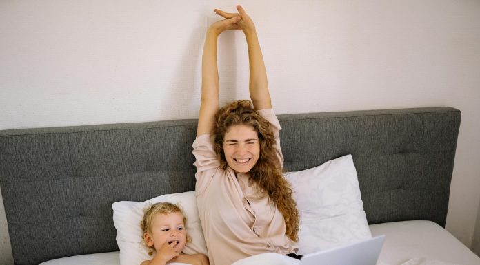 mother stretching in bed smiling while young child is next to her looking at her laptop; morning routine