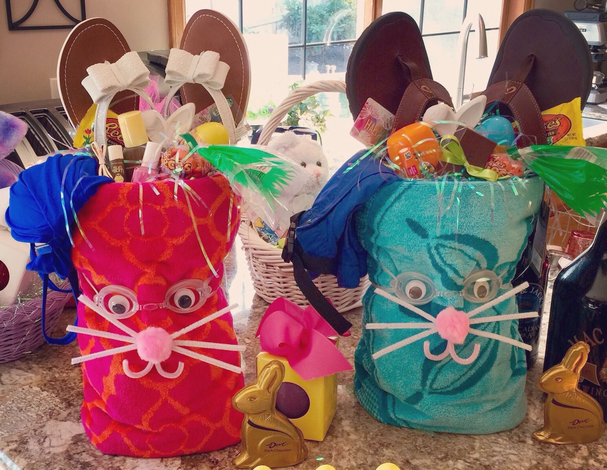 two Easter Baskets made out of rolled up beach towels with flip flops out of the top as ears on a counter with toys and chocolate bunnies in front