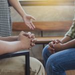 Two Christian people are praying for young man friends in church prayer room to encourage and support him in his problem and spiritual growth, small prayer group and fellowship concept.