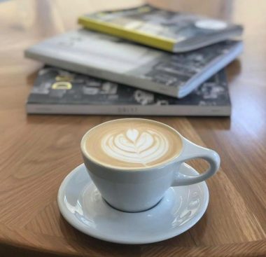 Latte art in a simple white coffee cup and saucer in front of a stack of books on top of a coffee table.