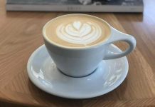 Latte art in a simple white coffee cup and saucer in front of a stack of books on top of a coffee table.