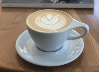 Latte art in a simple white coffee cup and saucer in front of a stack of books on top of a coffee table.