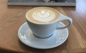 Latte art in a simple white coffee cup and saucer in front of a stack of books on top of a coffee table.