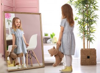 Cute little girl with teddy bear looking in mirror at home