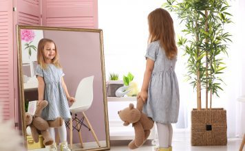 Cute little girl with teddy bear looking in mirror at home