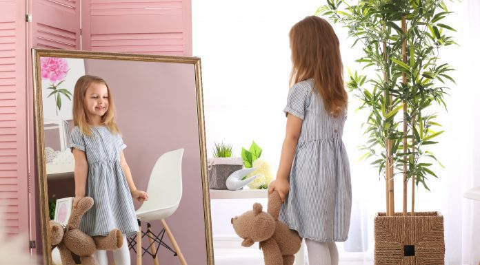 Cute little girl with teddy bear looking in mirror at home