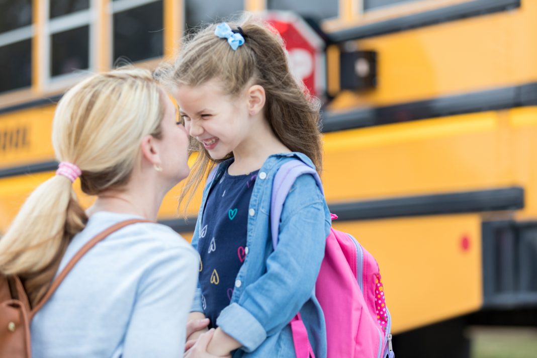 Mom gives daughter eskimo kiss before the girl boards school bus. The girl is excited about her first day of kindergarten. The school bus is in the background.