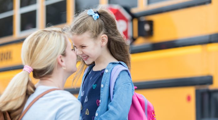 Mom gives daughter eskimo kiss before the girl boards school bus. The girl is excited about her first day of kindergarten. The school bus is in the background.