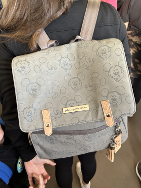 gray bag with beige accents with different faces of Mickey Mouse on it on the back of a woman in a black top with long brown hair facing away from camera