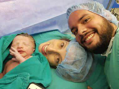 Mom just gave birth to baby via section. The baby is to the left of the picture with eyes closed, mom is smiling at picture and dad is to the right also smiling. 