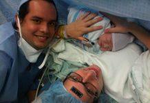 mom just having given birth via c-section to a baby boy. Mom is wearing a medical cap, with a black temperature sticker on her forehead, black glasses and holding baby. Dad is off to the left. Both are smiling.