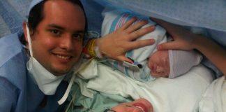 mom just having given birth via c-section to a baby boy. Mom is wearing a medical cap, with a black temperature sticker on her forehead, black glasses and holding baby. Dad is off to the left. Both are smiling.