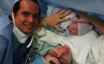 mom just having given birth via c-section to a baby boy. Mom is wearing a medical cap, with a black temperature sticker on her forehead, black glasses and holding baby. Dad is off to the left. Both are smiling.
