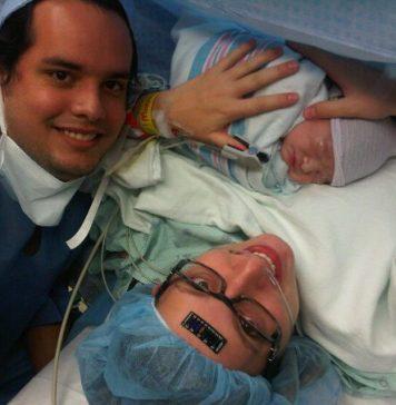 mom just having given birth via c-section to a baby boy. Mom is wearing a medical cap, with a black temperature sticker on her forehead, black glasses and holding baby. Dad is off to the left. Both are smiling.