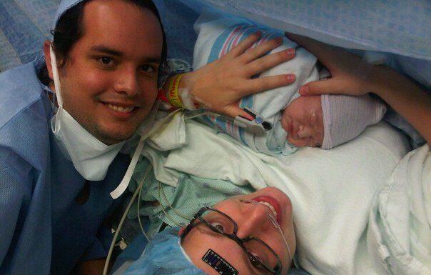 mom just having given birth via c-section to a baby boy. Mom is wearing a medical cap, with a black temperature sticker on her forehead, black glasses and holding baby. Dad is off to the left. Both are smiling.
