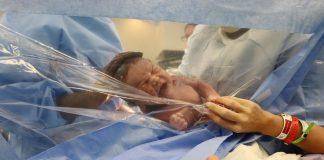 baby pushed against clear plastic after a c-section to show mama who reaches hand to clear plastic.