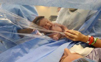 baby pushed against clear plastic after a c-section to show mama who reaches hand to clear plastic.