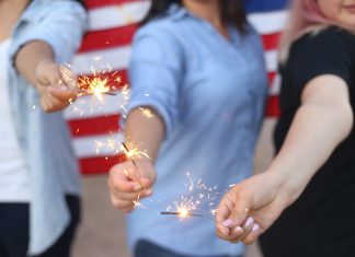 Alondra Olivas via Unsplash three women hold sparklers with an American flag behind them