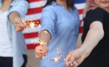 Alondra Olivas via Unsplash three women hold sparklers with an American flag behind them
