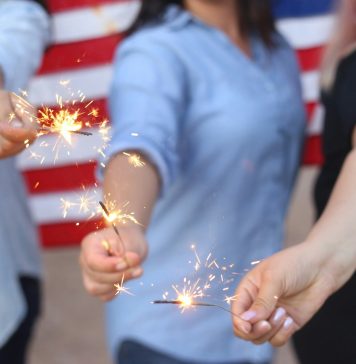 Alondra Olivas via Unsplash three women hold sparklers with an American flag behind them