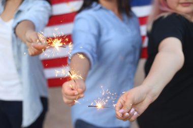 Alondra Olivas via Unsplash three women hold sparklers with an American flag behind them