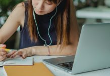 girl writing in notebook with earbuds and a computer in front of her, going through college asmission