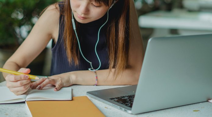 girl writing in notebook with earbuds and a computer in front of her, going through college asmission