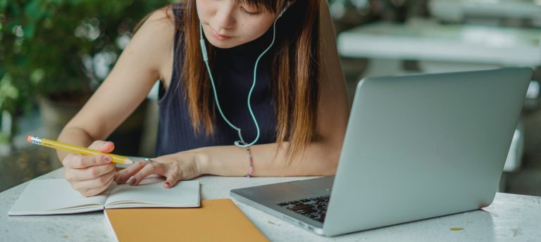 girl writing in notebook with earbuds and a computer in front of her, going through college asmission