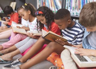 Side view of mixed ethnicity school kids sitting on cushions and studying over books in a library at school against bookshelfs in background; black history month