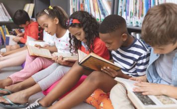 Side view of mixed ethnicity school kids sitting on cushions and studying over books in a library at school against bookshelfs in background; black history month