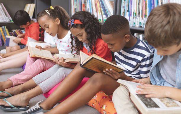 Side view of mixed ethnicity school kids sitting on cushions and studying over books in a library at school against bookshelfs in background; black history month