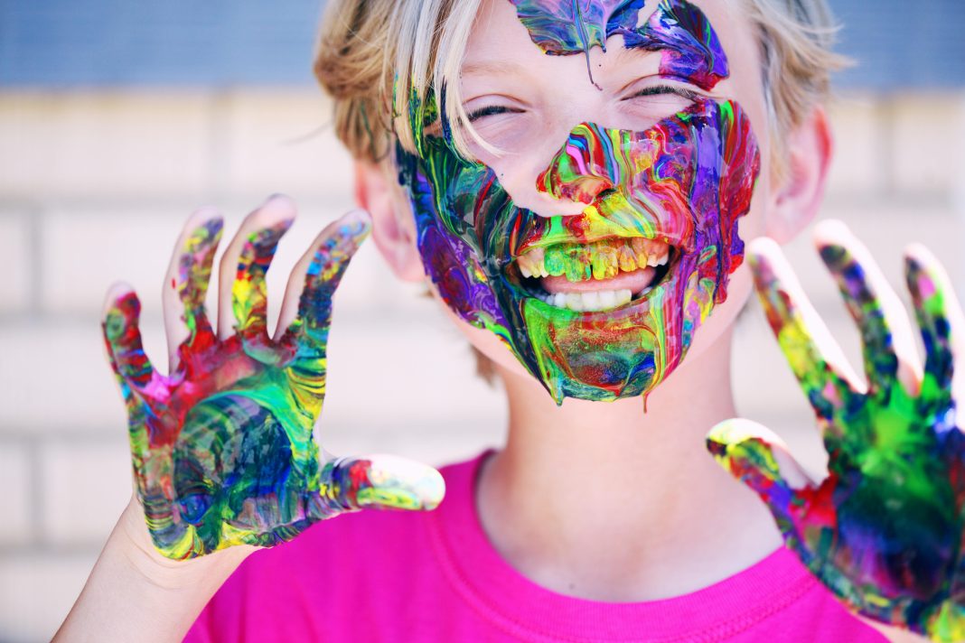 child laughing at camera with colorful paint on hands and all over face, educational resources