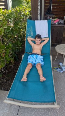 young boy sitting in a lounge chair in a bathing suit staring at the camera smiling