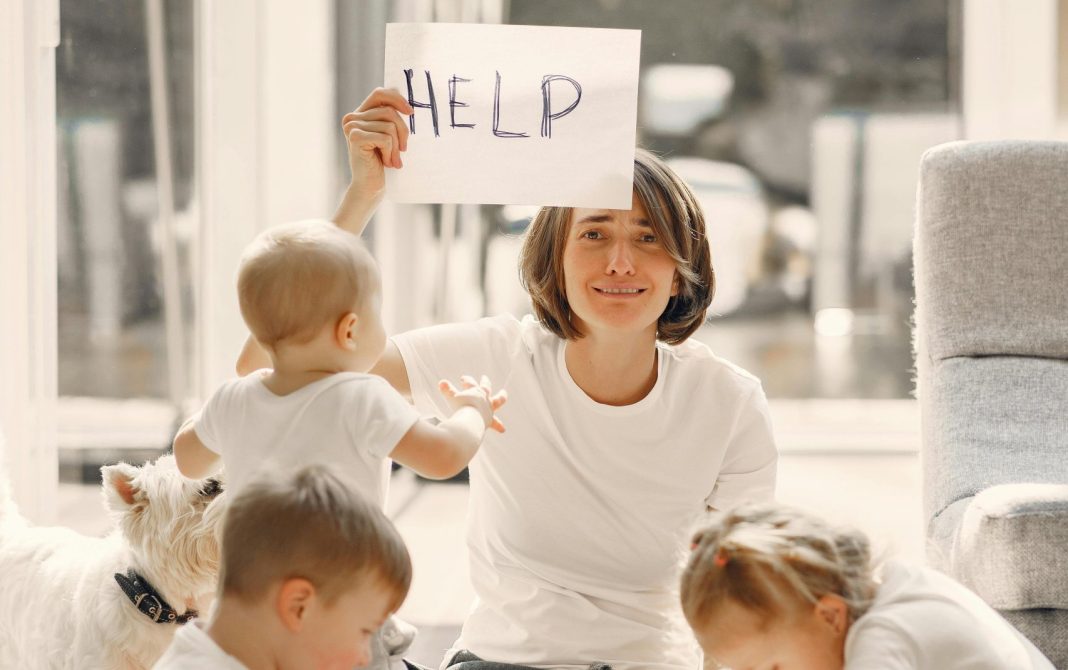 mom holding up a sign that says "help," with children around on her with school supplies for back to school madness