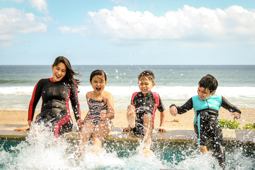 A mom and her three children smiling and splashing water wearing wetsuits sitting on a dock.