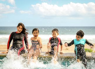 A mom and her three children smiling and splashing water wearing wetsuits sitting on a dock.