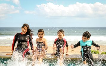 A mom and her three children smiling and splashing water wearing wetsuits sitting on a dock.