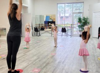 a teacher with a group of young girls in ballerina uniforms with the arms up in a dance class discussing the benefits of dance