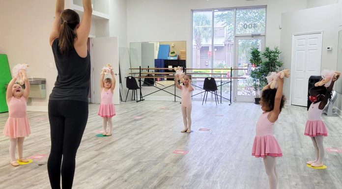 a teacher with a group of young girls in ballerina uniforms with the arms up in a dance class discussing the benefits of dance