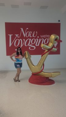 woman standing next to a large golden anchor with a sign that says "now we're voyaging". Woman is smiling at the camera