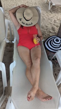 woman laying down on lounge chair in bathing suit with a drink in her hand, hat covering her face hat says "do not disturb"